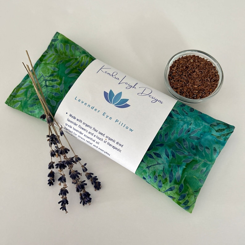 May include: A green and blue patterned lavender eye pillow with a white label that reads "Kendra Leigh Designs Lavender Eye Pillow." A small bowl of seeds and a bundle of dried lavender are next to the pillow.
