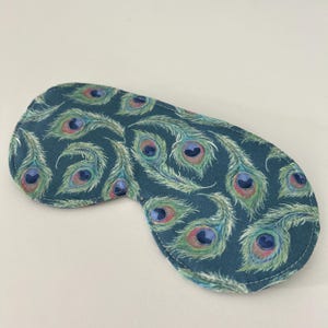 May include: A teal sleep mask with a repeating pattern of peacock feathers in shades of blue, green, pink, and purple. The mask is oval-shaped with a white stitched border. The sleep mask is designed to block out light.