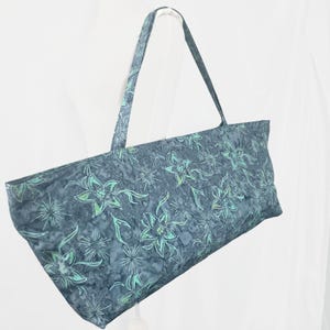 May include: A blue yoga mat bag featuring a floral pattern in teal and white. The bag has a rectangular shape with a long handle. The floral design is repeated across the fabric, suitable for carrying a yoga mat.
