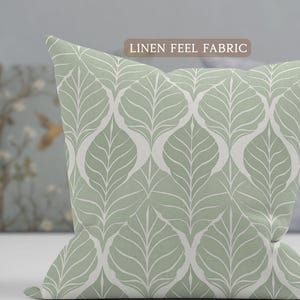 May include: A square decorative pillow with a light green background and a white leaf pattern. The pillow has a linen feel fabric. The pillow is on a white surface.