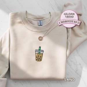 Gildan 18000 sand embroidered sweatshirt featuring a small boba tea cup with black tapioca pearls and green straw stitched on the chest, shown folded flat on a marble background