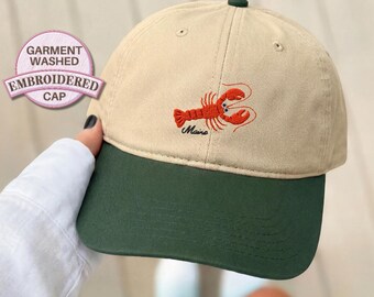 Könnte beinhalten: Khaki- und dunkelgrüne Baseballkappe mit gesticktem rotem Hummer-Design. Unter dem Hummer ist der Schriftzug "Maine" gestickt. Die Kappe trägt auch den Text "Garment Washed Embroidered Cap".