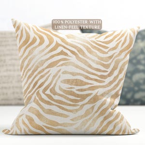 May include: A square throw cushion featuring a beige and white zebra stripe pattern. The fabric is 100% polyester with a linen-feel texture. The design consists of organic, wavy lines in a light tan colour against a white background, creating a repeating animal print motif across the entire surface of the cushion cover.