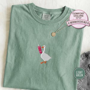 Light green embroidered T-shirt with a small goose holding a pink drink cup stitched on the chest, shown folded with denim jeans for a playful casual outfit