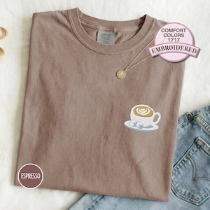 Comfort Colors 1717 espresso embroidered T-shirt featuring a latte coffee cup design with sugar cubes, shown folded flat with denim jeans for a cozy café-inspired casual outfit