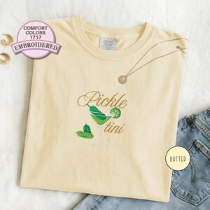 Comfort Colors 1717 butter color embroidered T-shirt featuring a pickle martini design with green olives and a cocktail glass, shown folded flat with denim jeans for a playful foodie aesthetic look