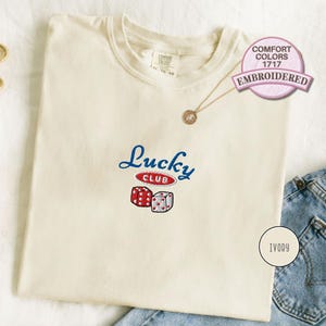 Könnte beinhalten: Cremefarbenes T-Shirt mit blau-roter "Lucky Club" Stickerei. Mit zwei Würfeln und dem Text "COMFORT COLORS 1717 EMBROIDERED". Eine goldene Halskette und Ohrringe sind ebenfalls zu sehen.