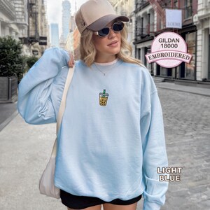 May include: A light blue crewneck sweatshirt featuring a small embroidered graphic of a bubble tea drink on the chest. The garment is labelled as a Gildan 18000 model. The sweatshirt has long sleeves and a relaxed fit, shown in a light blue colour.