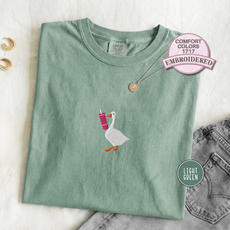 Light green embroidered T-shirt with a small goose holding a pink drink cup stitched on the chest, shown folded with denim jeans for a playful casual outfit