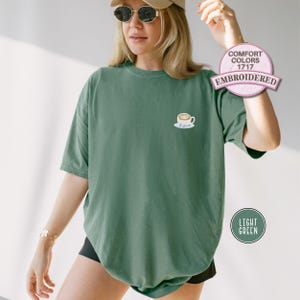 Puede incluir: Camiseta verde claro con un peque&ntilde;o dise&ntilde;o de taza de caf&eacute; bordada. La camiseta tiene mangas cortas y un corte holgado. El texto "COMFORT COLORS 1717 EMBROIDERED" est&aacute; en un c&iacute;rculo rosa. El texto "LIGHT GREEN" est&aacute; en un c&iacute;rculo verde.