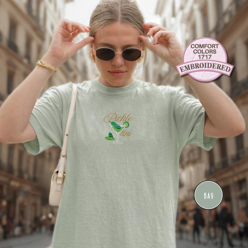 May include: A light green t-shirt with the embroidered text "Pickle Martini" and a matching graphic of a martini glass. The shirt has short sleeves and a crew neck. The shirt is labeled "Comfort Colors 1717 Embroidered".