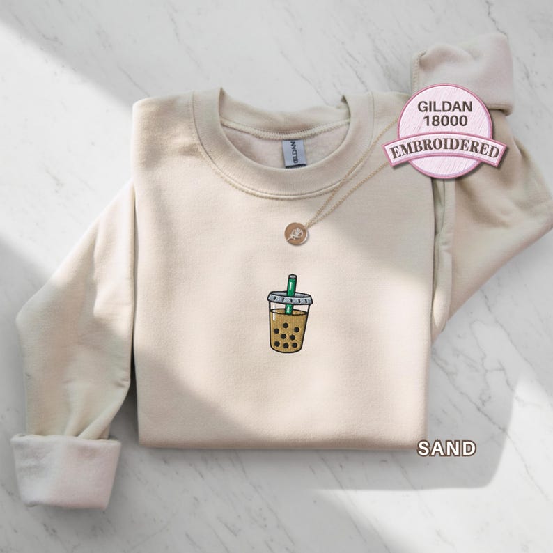Gildan 18000 sand embroidered sweatshirt featuring a small boba tea cup with black tapioca pearls and green straw stitched on the chest, shown folded flat on a marble background