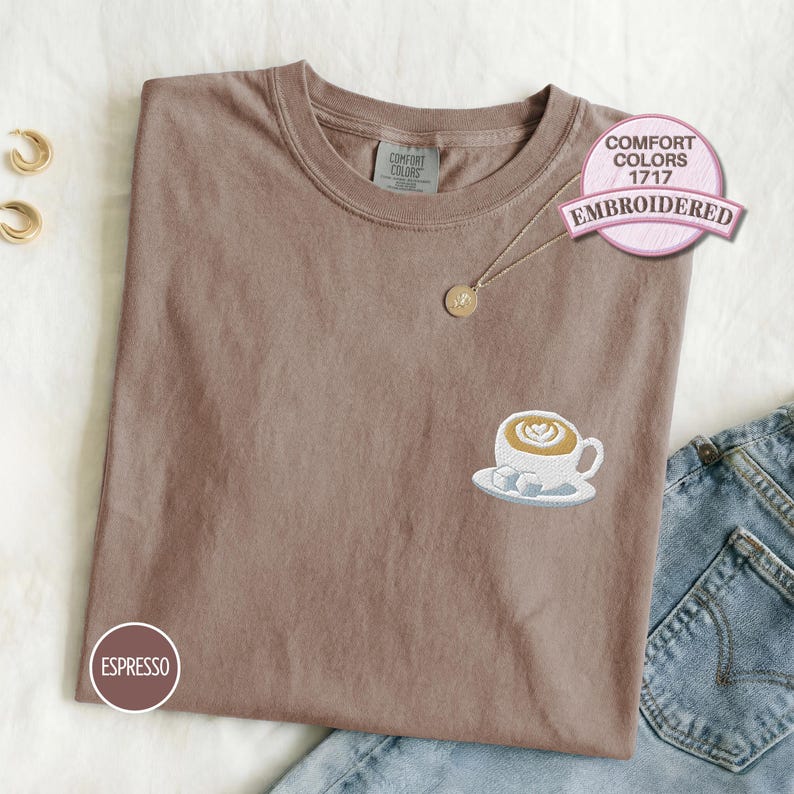 Comfort Colors 1717 espresso embroidered T-shirt featuring a latte coffee cup design with sugar cubes, shown folded flat with denim jeans for a cozy café-inspired casual outfit