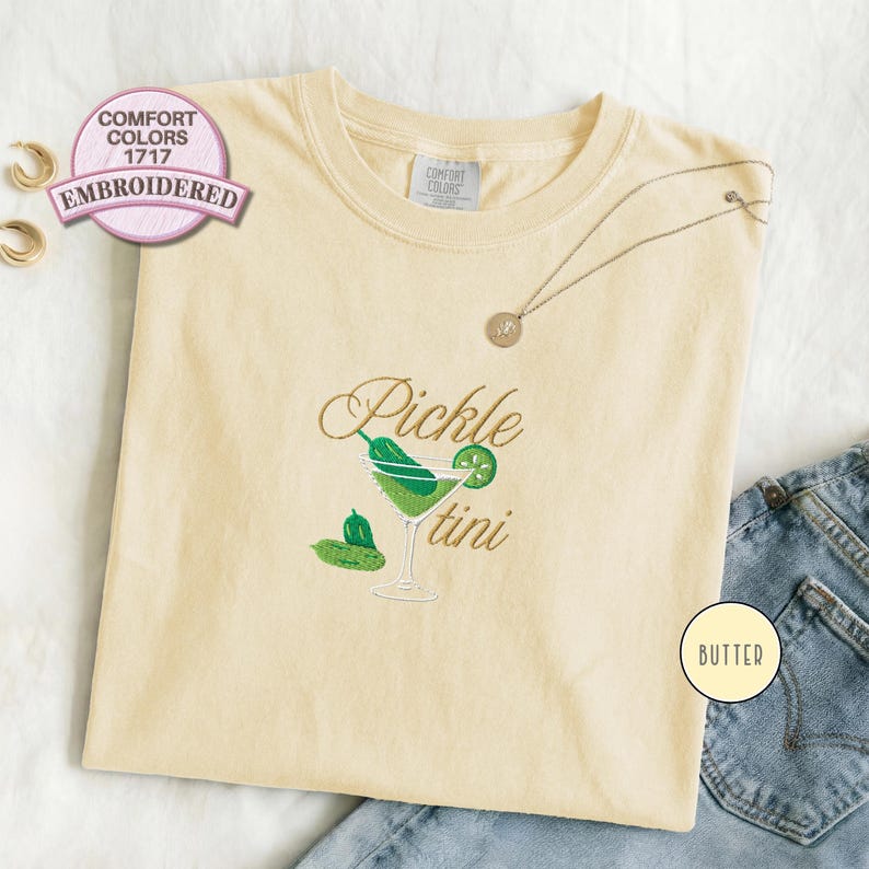 Comfort Colors 1717 butter color embroidered T-shirt featuring a pickle martini design with green olives and a cocktail glass, shown folded flat with denim jeans for a playful foodie aesthetic look