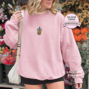 May include: A light pink crewneck sweatshirt featuring a small embroidered graphic of a bubble tea cup with a green straw on the chest. The garment includes a circular badge stating GILDAN 18000 EMBROIDERED and a label indicating LIGHT PINK.