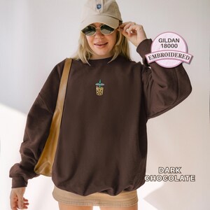 May include: A dark chocolate brown crewneck sweatshirt featuring a small embroidered graphic of a boba tea drink on the centre chest. The garment is labelled as Gildan 18000 and is shown in the colour dark chocolate with long sleeves and a ribbed hem.