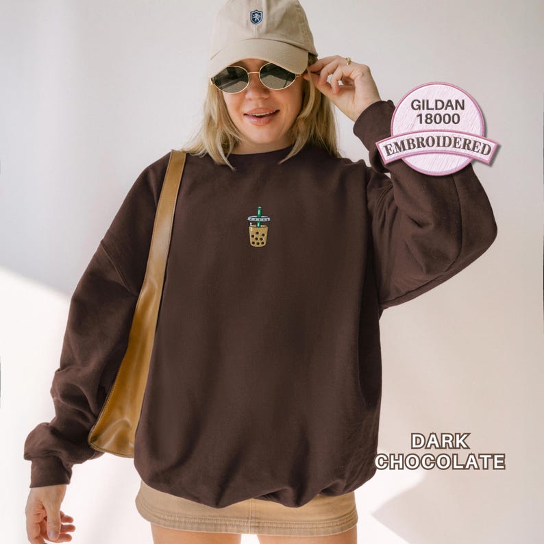 May include: A dark chocolate brown crewneck sweatshirt featuring a small embroidered graphic of a boba tea drink on the centre chest. The garment is labelled as Gildan 18000 and is shown in the colour dark chocolate with long sleeves and a ribbed hem.