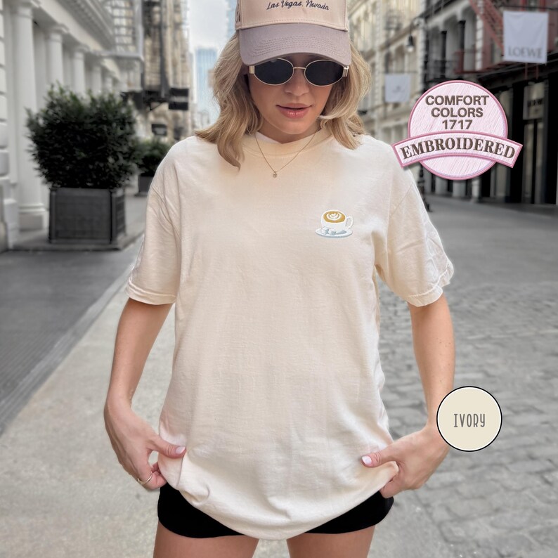 Puede incluir: Camiseta color marfil con un dise&ntilde;o de taza de caf&eacute; bordada. La camiseta tiene mangas cortas y cuello redondo. La imagen tambi&eacute;n muestra una gorra beige y pantalones cortos negros. La camiseta est&aacute; etiquetada como "Comfort Colors 1717 Embroidered" e "Ivory".