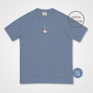 May include: Blue jean colored t-shirt with a small embroidered design of a goose holding a pink drink. The shirt is made of 100% cotton. The shirt has a classic crew neck and short sleeves.