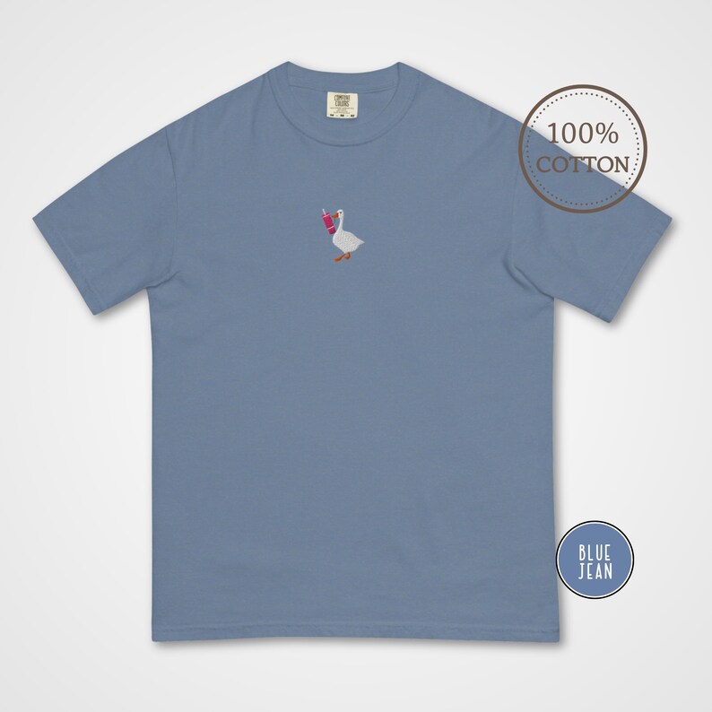 May include: Blue jean colored t-shirt with a small embroidered design of a goose holding a pink drink. The shirt is made of 100% cotton. The shirt has a classic crew neck and short sleeves.