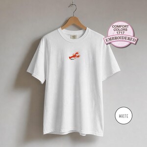 May include: A white short-sleeve t-shirt featuring a small embroidered red lobster graphic on the center chest with the word "Mama" stitched below it. The shirt is displayed on a wooden hanger and includes a circular badge labeled COMFORT COLORS 1717 EMBROIDERED.