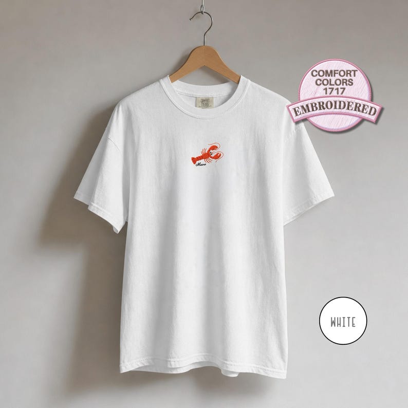 May include: A white short-sleeve t-shirt featuring a small embroidered red lobster graphic on the center chest with the word "Mama" stitched below it. The shirt is displayed on a wooden hanger and includes a circular badge labeled COMFORT COLORS 1717 EMBROIDERED.
