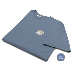 Puede incluir: Una camiseta azul vaquero con un peque&ntilde;o dise&ntilde;o de taza de caf&eacute; bordada en el pecho. La camiseta est&aacute; doblada y tiene una etiqueta en el cuello. Una pegatina circular con las palabras "BLUE JEAN" est&aacute; en la esquina inferior derecha.