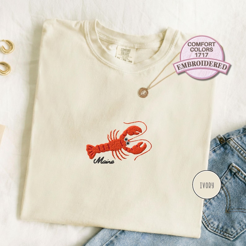 Ivory Comfort Colors 1717 T-shirt featuring an embroidered red lobster with raised claws, curved antennae, and black Maine lettering, styled flat with denim and gold jewelry.