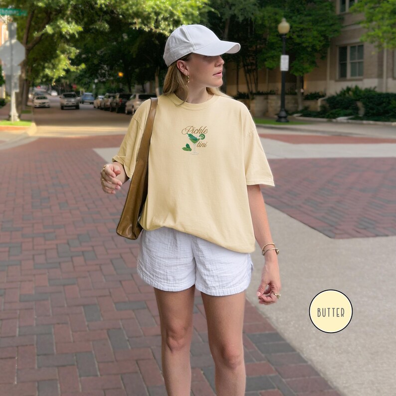 May include: A light yellow t-shirt with the text "Pickle tini" in green and brown. The shirt is worn with white shorts and a white baseball cap. A brown shoulder bag is also visible.