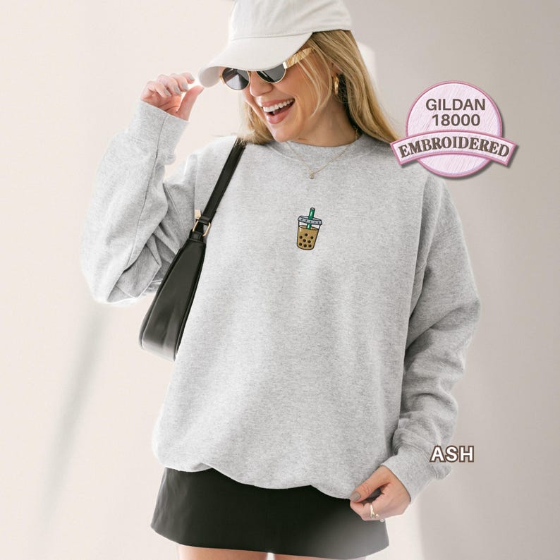 May include: A light grey crewneck sweatshirt featuring a small embroidered graphic of a bubble tea drink with a green straw on the chest. The sweatshirt has long sleeves and a ribbed neckline. A black shoulder bag is worn over one side.