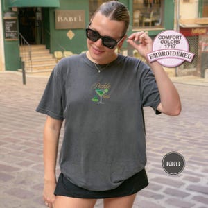 May include: Dark gray t-shirt with the embroidered text "Pickle Martini" and a green cocktail graphic. The shirt is a Comfort Colors 1717 style in the color Pepper.
