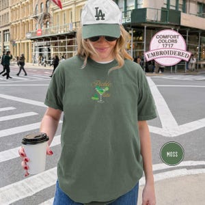 May include: Moss green t-shirt with a "Pickle Martini" design embroidered on the front. The design features a martini glass and a pickle. The shirt is a Comfort Colors 1717, embroidered.