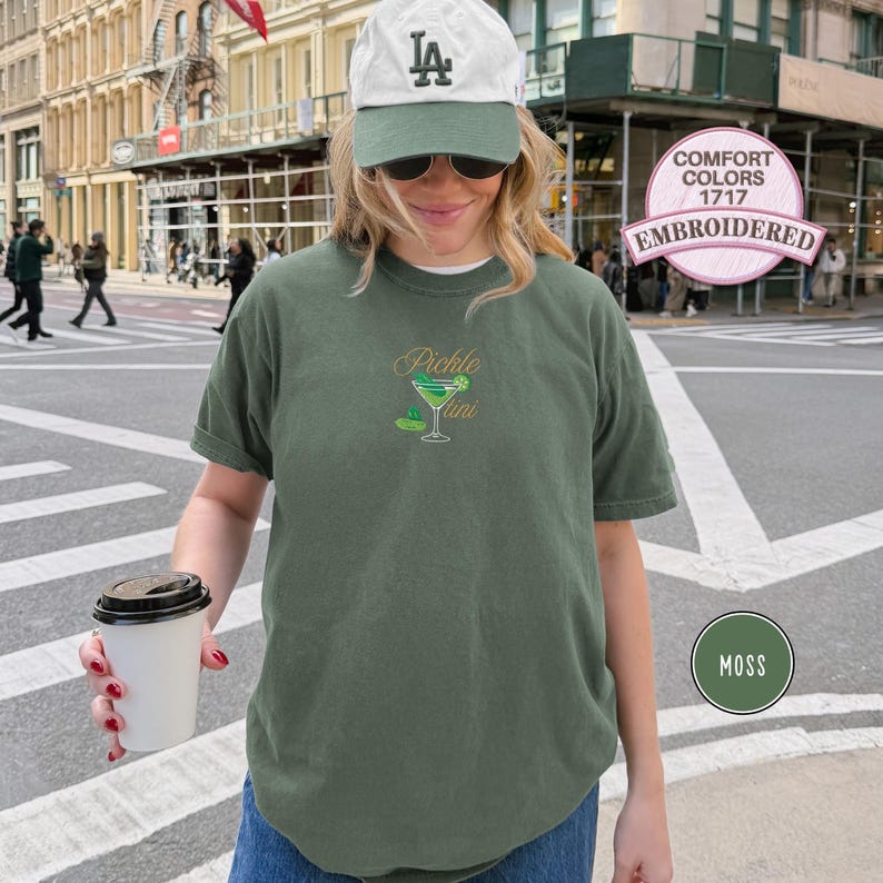 May include: Moss green t-shirt with a "Pickle Martini" design embroidered on the front. The design features a martini glass and a pickle. The shirt is a Comfort Colors 1717, embroidered.