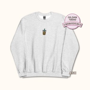 May include: An ash grey crewneck sweatshirt featuring a small, centred embroidered graphic of a brown bubble tea drink with a green straw. The garment includes a Gildan 18000 label and is made of a heathered fabric with long sleeves and ribbed cuffs.