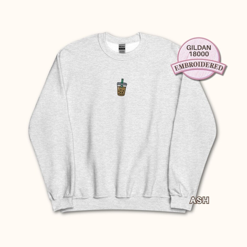 May include: An ash grey crewneck sweatshirt featuring a small, centred embroidered graphic of a brown bubble tea drink with a green straw. The garment includes a Gildan 18000 label and is made of a heathered fabric with long sleeves and ribbed cuffs.