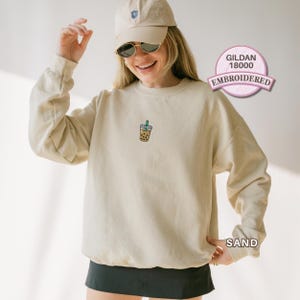 May include: A sand-coloured crewneck sweatshirt featuring a small embroidered graphic of a bubble tea drink with a green straw on the centre chest. The garment is a Gildan 18000 model, shown in a light beige tone, paired with a matching baseball cap.