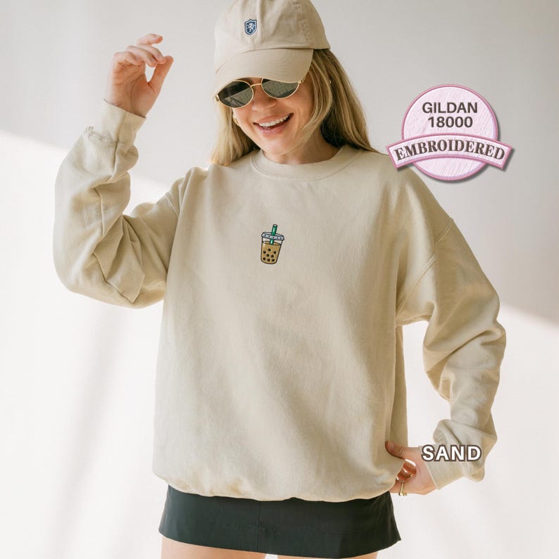 May include: A sand-coloured crewneck sweatshirt featuring a small embroidered graphic of a bubble tea drink with a green straw on the centre chest. The garment is a Gildan 18000 model, shown in a light beige tone, paired with a matching baseball cap.