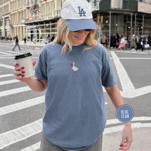 May include: Blue jean colored short-sleeve t-shirt with an embroidered goose design holding a pink and red object. The shirt is worn with a blue baseball cap and sunglasses. A coffee cup is held in the hand.