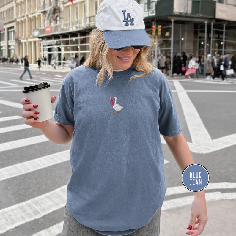 May include: Blue jean colored short-sleeve t-shirt with an embroidered goose design holding a pink and red object. The shirt is worn with a blue baseball cap and sunglasses. A coffee cup is held in the hand.