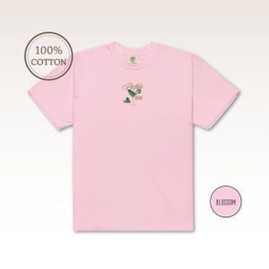 May include: A light pink t-shirt with the embroidered text "Pickle tini" and a martini glass design with a pickle and olive. The shirt is made of 100% cotton and has a round neckline. The word "Blossom" is in a pink circle.