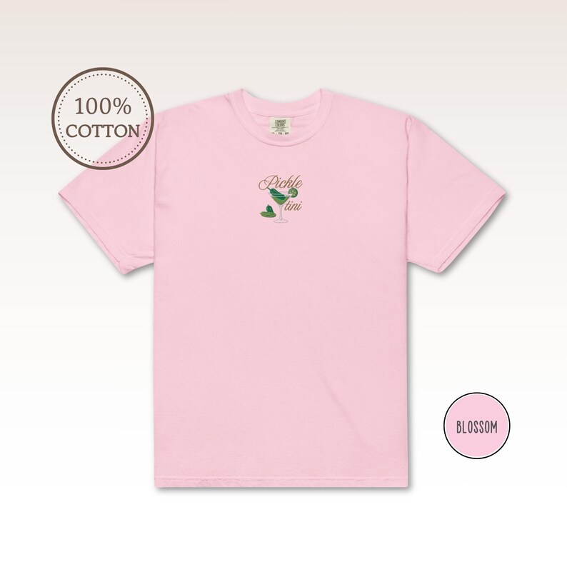 May include: A light pink t-shirt with the embroidered text "Pickle tini" and a martini glass design with a pickle and olive. The shirt is made of 100% cotton and has a round neckline. The word "Blossom" is in a pink circle.