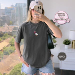 May include: Pepper-colored Comfort Colors 1717 embroidered t-shirt featuring a small embroidered goose with a pink bow. The shirt is a short-sleeved, crew neck style. The image also shows a beige cap and denim shorts.
