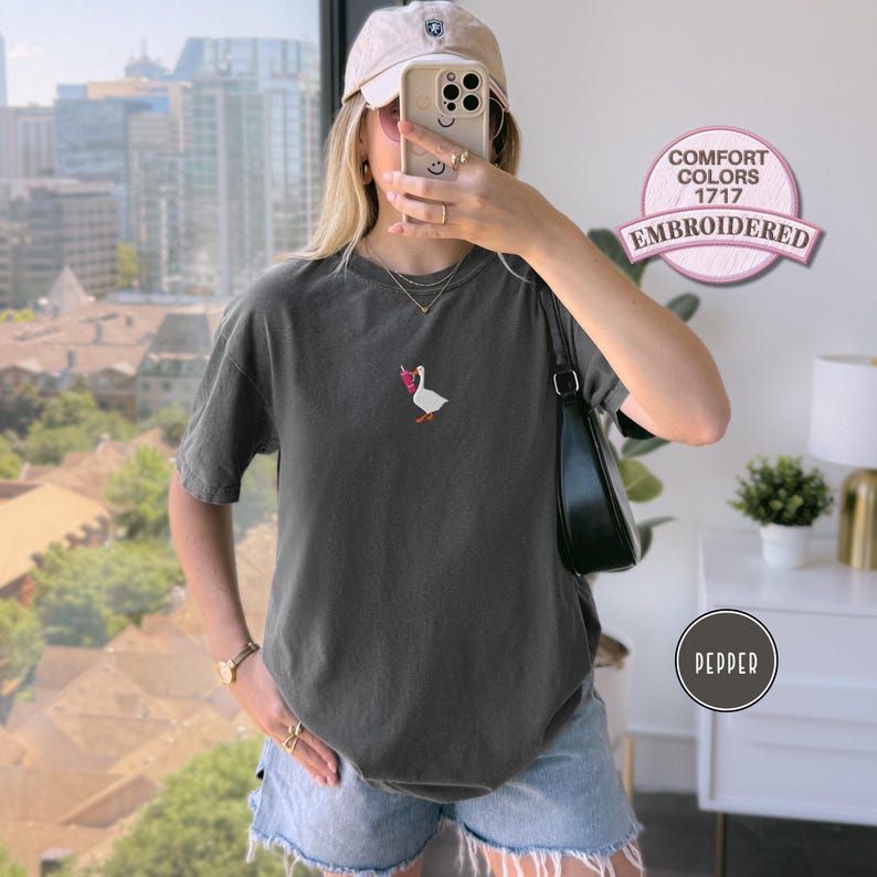 May include: Pepper-colored Comfort Colors 1717 embroidered t-shirt featuring a small embroidered goose with a pink bow. The shirt is a short-sleeved, crew neck style. The image also shows a beige cap and denim shorts.