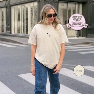 May include: Ivory-colored t-shirt with an embroidered goose design. The shirt is a Comfort Colors 1717 style. The design features a goose with a red bow. The shirt is paired with blue jeans.