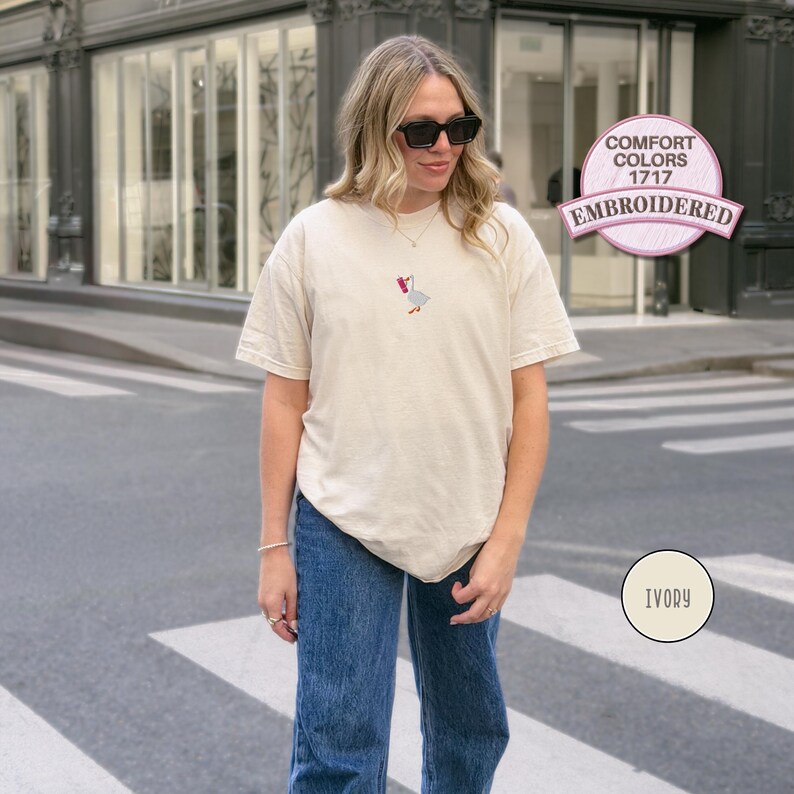 May include: Ivory-colored t-shirt with an embroidered goose design. The shirt is a Comfort Colors 1717 style. The design features a goose with a red bow. The shirt is paired with blue jeans.