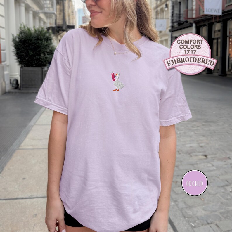 May include: Orchid-colored t-shirt with an embroidered goose holding a red drink. The shirt is labeled "Comfort Colors 1717 Embroidered". A small, round "Orchid" label is also visible. The shirt is a short-sleeved, casual style.
