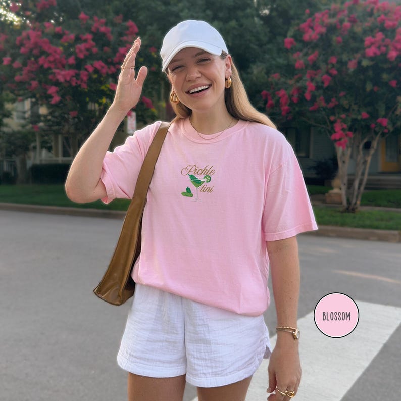 May include: Pink t-shirt with the embroidered text "Pickle Martini" and a cocktail graphic. The shirt is paired with white shorts and a white baseball cap. A brown shoulder bag is also visible.