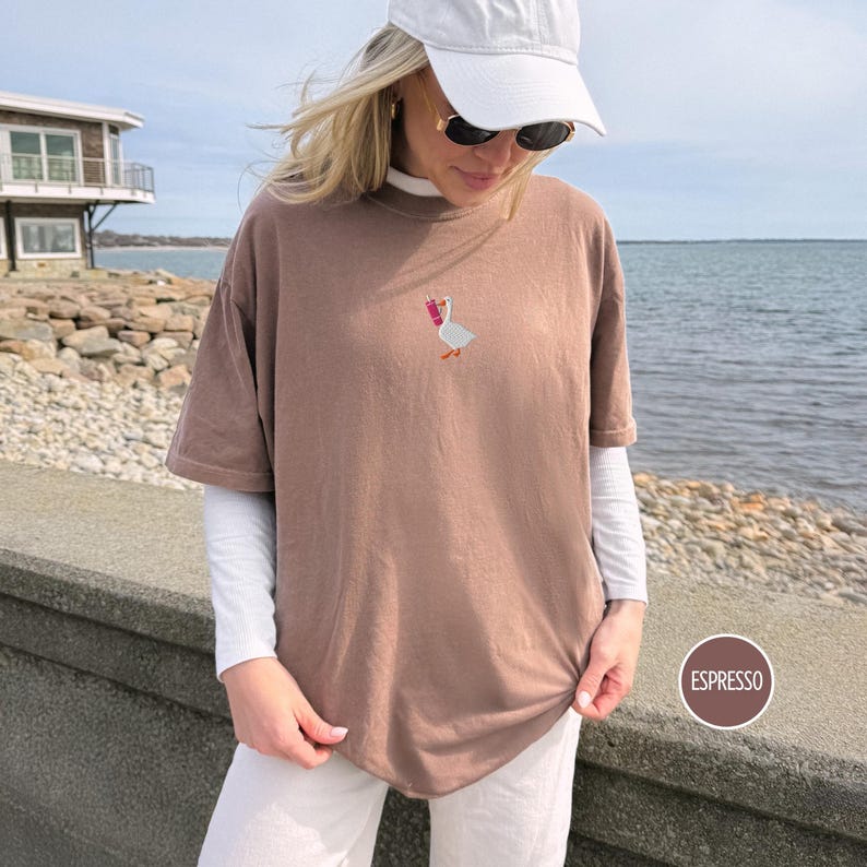 May include: Espresso-colored t-shirt with a small embroidered goose holding a pink drink. The shirt has short sleeves and a relaxed fit. The model is wearing a white baseball cap, sunglasses, and a white long-sleeved shirt underneath. The word "Espresso" is in a brown circle.