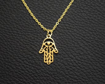 May include: A gold-coloured necklace featuring a Hamsa hand pendant. The pendant has an eye in the centre and detailed openwork patterns. The chain is also gold, set against a black, textured backdrop.