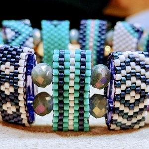 May include: A beaded bracelet with a geometric pattern in shades of blue, white, and green. The bracelet is made with small, square beads and features iridescent accents.